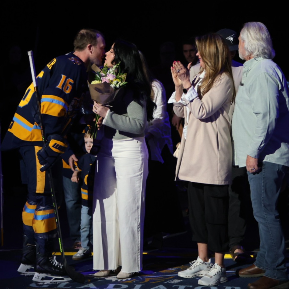 A hockey player in a team jersey kisses a woman holding a bouquet of flowers, while family members stand nearby, expressing joy during a special event.