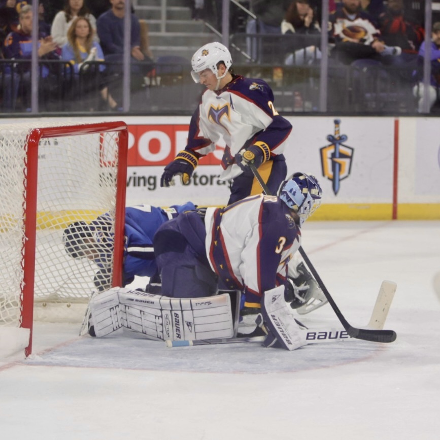 An ice hockey player in a white jersey is about to jump over a fallen goaltender in blue gear, who is on the ice behind the net.