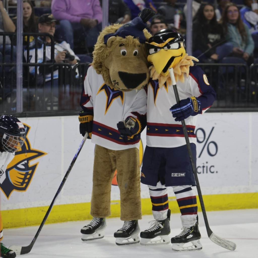 Two sports mascots, a lion and a bird, pose together on the ice during a hockey game, with a young player in the background.