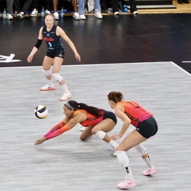 A volleyball player in an orange jersey dives to save the ball during a match, while another player in pink prepares for the play and a third player looks on from the background.
