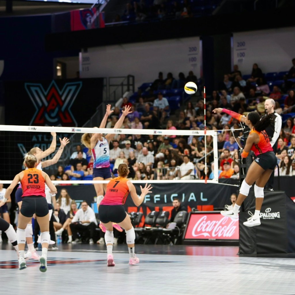 A volleyball match in action with players jumping and reaching for the ball over the net. The player in mid-air is about to spike the ball, while teammates prepare to react. The crowd is visible in the background.