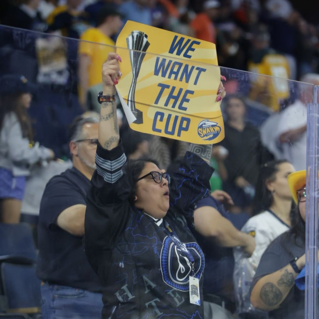 A passionate fan holding a sign that reads 'WE WANT THE CUP' while cheering in a crowded sports arena.