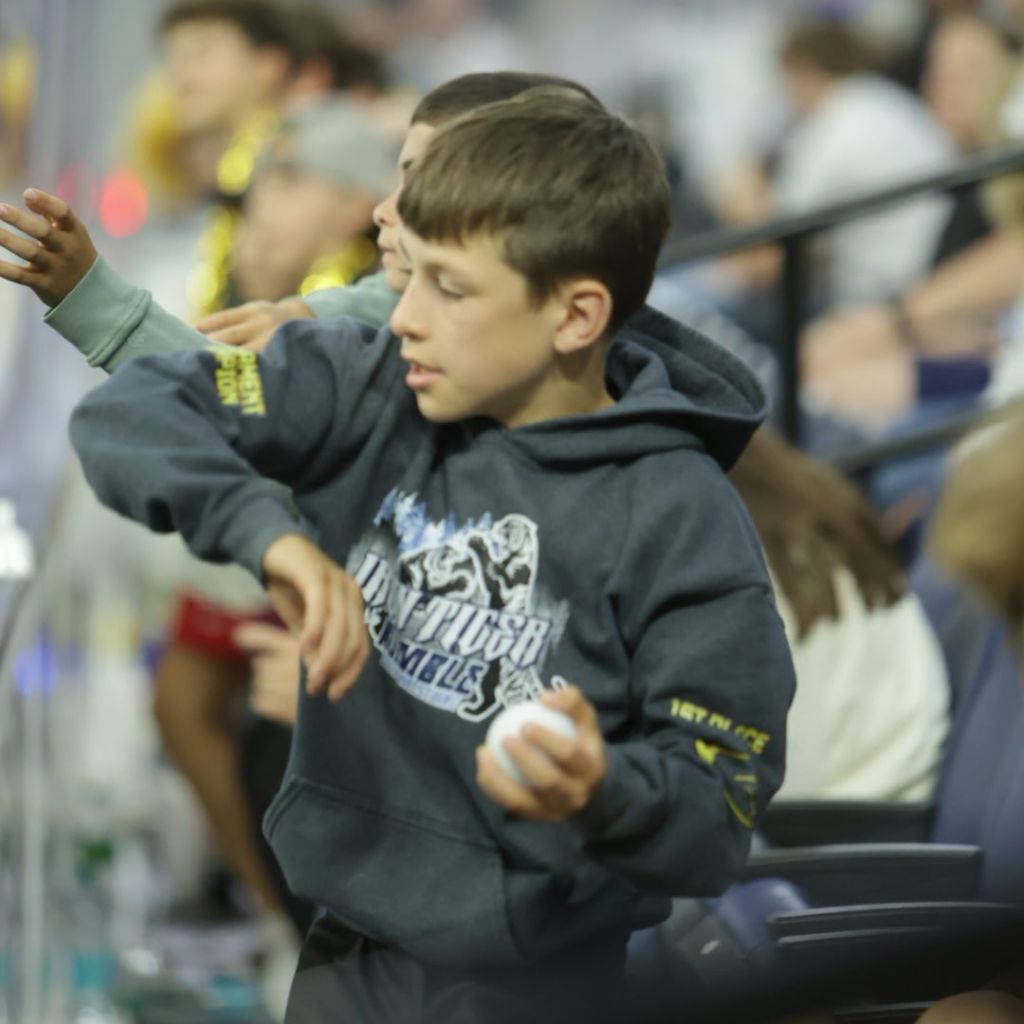 A young boy wearing a dark hoodie is focused on something in front of him while holding a small white ball. Other children are partially visible in the background.