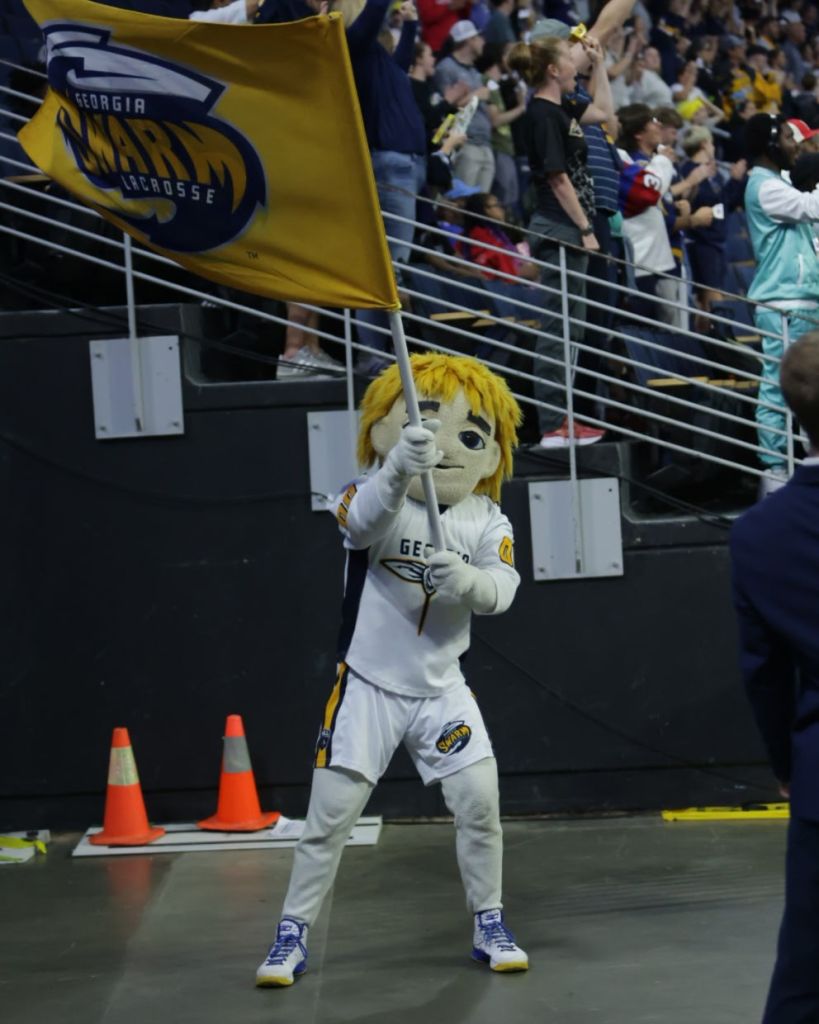 A mascot wearing a sports uniform and a blonde wig, holding a flag with the 'Georgia Swarm Lacrosse' logo, cheerfully engaging with the crowd at a sporting event.