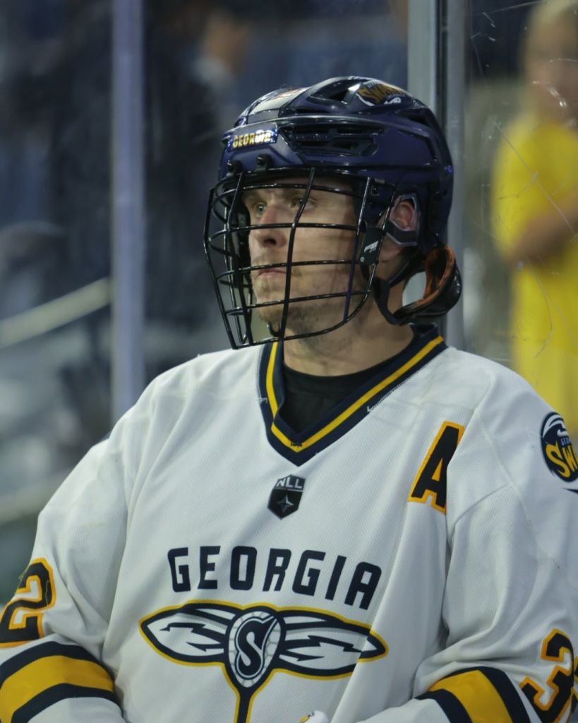 A lacrosse player wearing a white jersey with 'Georgia' printed on it, staring thoughtfully while standing by the glass during a game.