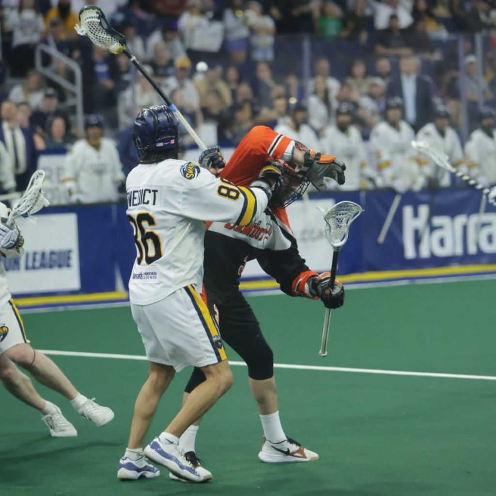 A tense moment during a lacrosse game showing players in action on the field. One player in white jersey is defending against an opponent in orange and black as they engage for control of the ball.