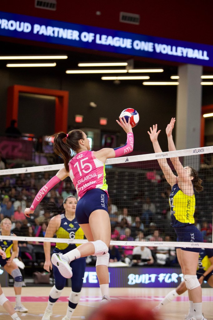 A volleyball player jumps to spike the ball over the net during a match, with two opposing players attempting to block. The setting is a sports arena with spectators in the background.