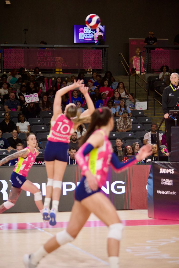 A volleyball player wearing a pink jersey jumps to serve a volleyball during a match, with spectators in the background cheering and holding signs.