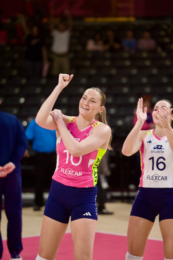 Two female volleyball players celebrating on the court, one in a pink jersey with the number 10 and the other in a white jersey with the number 16.