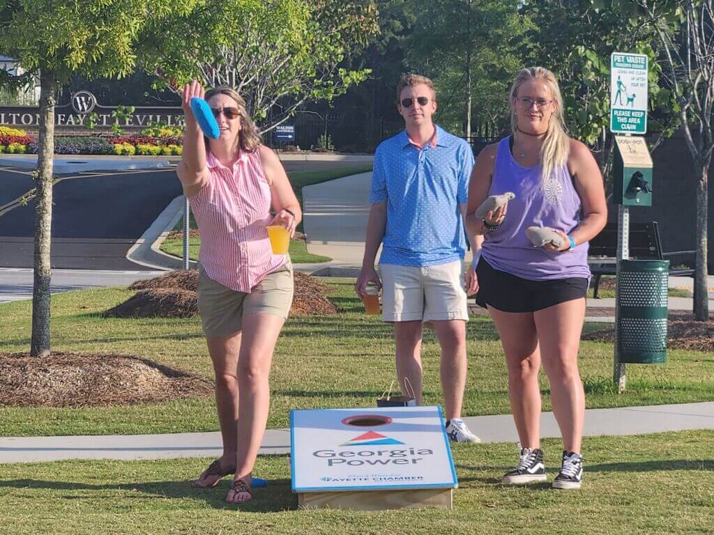 A woman throws a blue bean bag while holding a drink, with two friends standing nearby in a park setting. They are playing a bean bag toss game with a 'Georgia Power' board.