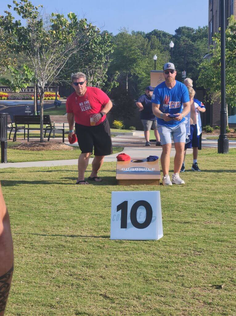 Two men playing cornhole outdoors, with one wearing a red shirt and the other in blue, aiming to throw bags towards the target board. A score display showing '10' is in the foreground.