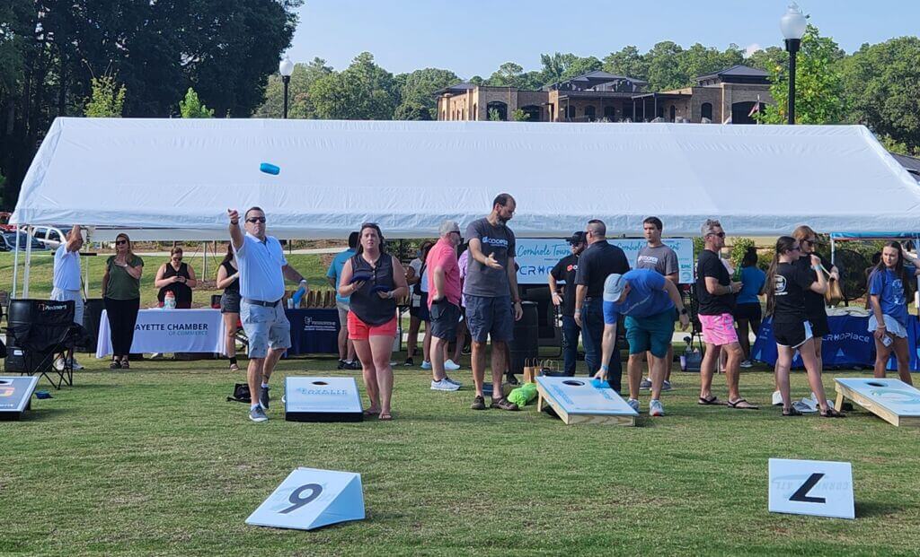 A lively outdoor scene at a community event featuring people playing cornhole under a large white tent. Participants are engaged in throwing bags towards cornhole boards, with banners and tables from local organizations in the background.