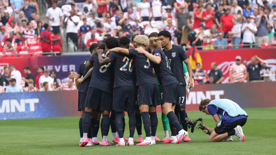 A group of soccer players in dark jerseys huddling together on the field, with fans in the background cheering.