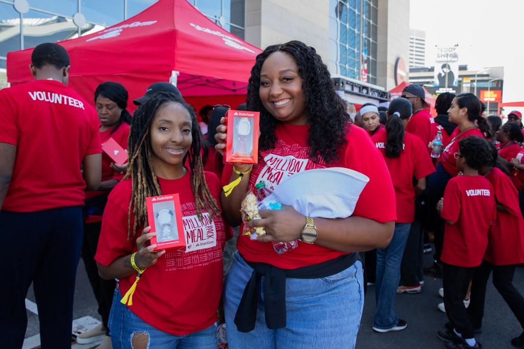 Two smiling women hold promotional items while surrounded by a group of volunteers in red shirts at an outdoor event.