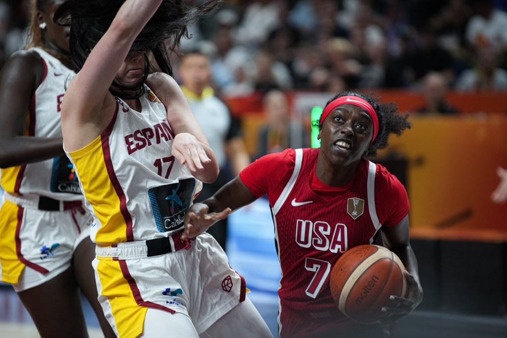 A basketball player wearing a red jersey with 'USA' on it is in action, attempting to score while being closely guarded by an opponent in a white jersey marked 'ESPAÑA'.