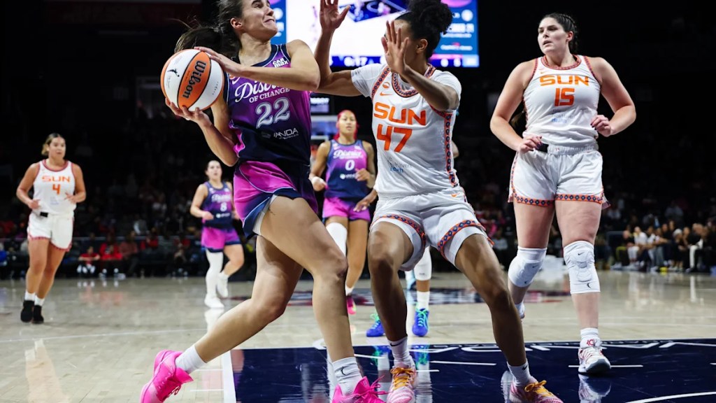 A basketball player in a purple uniform attempts to make a play while being guarded by a player in a white uniform with 'SUN' on it. Other players are visible in the background, and the game is taking place in a brightly lit arena.