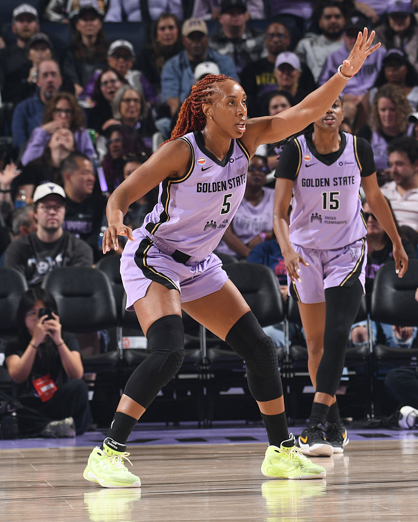 A female basketball player in a purple uniform, number 5, is in motion on the court, communicating with her teammate, while a crowd is visible in the background.