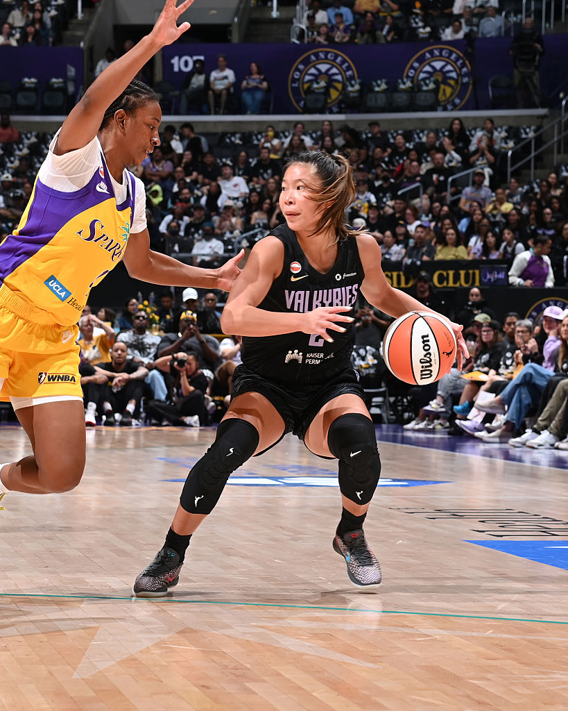 A female basketball player in a black jersey dribbles the ball while being defended by an opponent in a purple and white jersey during a WNBA game. The scene captures the intensity of the match, with spectators visible in the background.