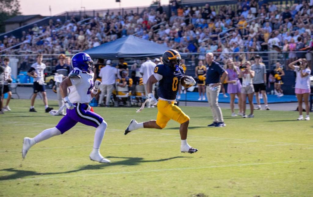 A football player in a dark blue jersey runs down the field while being chased by an opponent in a white and purple uniform during a high school football game.