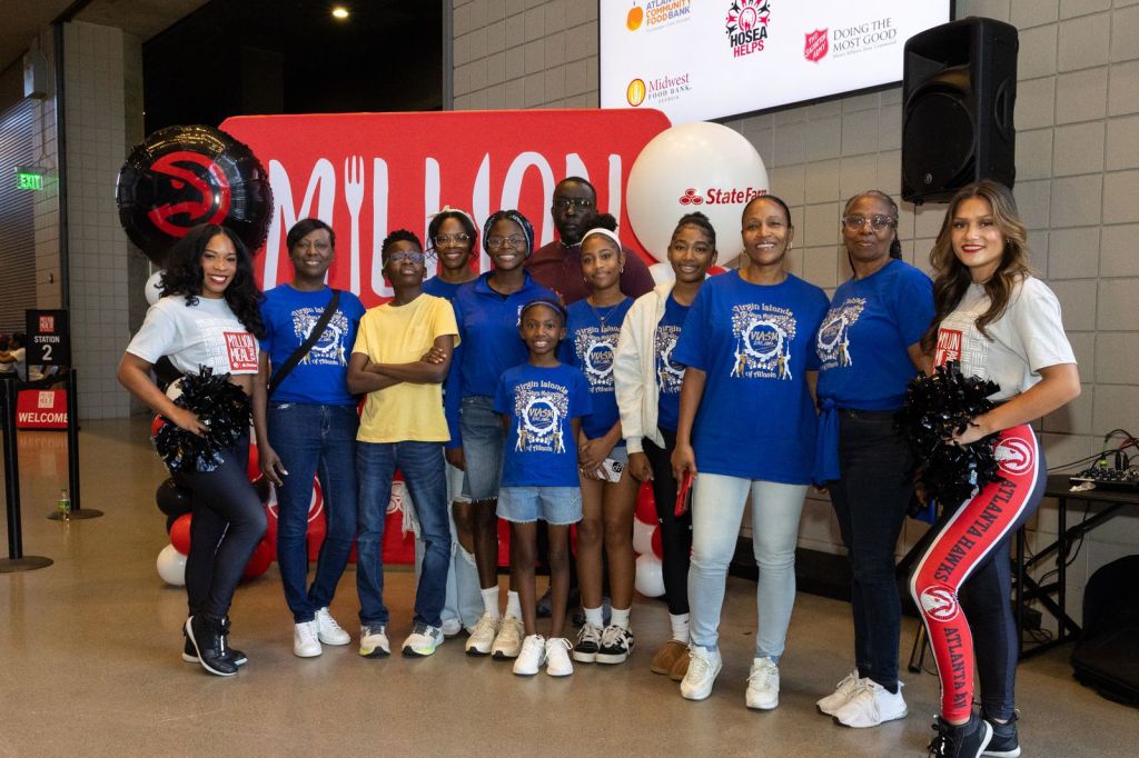 A group of people posing together at an event, wearing blue shirts with a design, alongside a woman in a cheerleading outfit. The background features promotional banners and balloons.