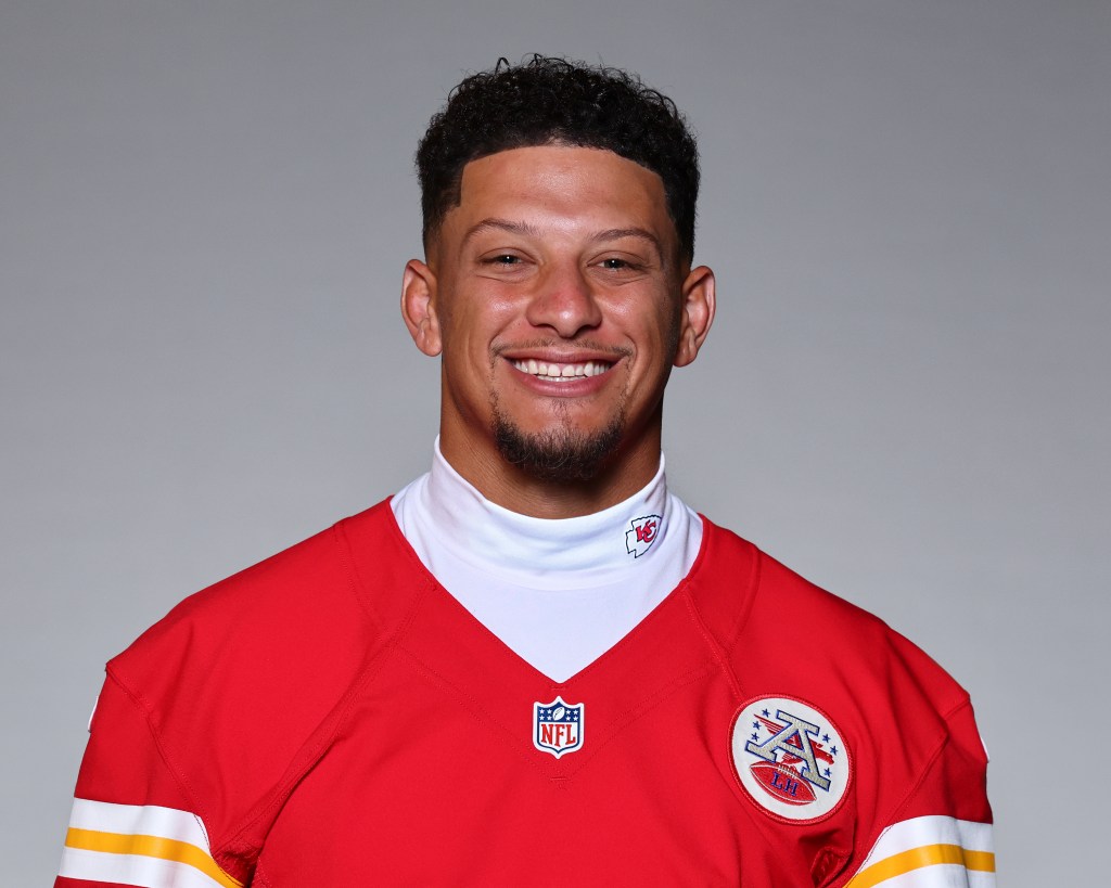 Close-up portrait of a smiling man wearing a red football jersey with white and yellow accents and an NFL logo, against a gray background.