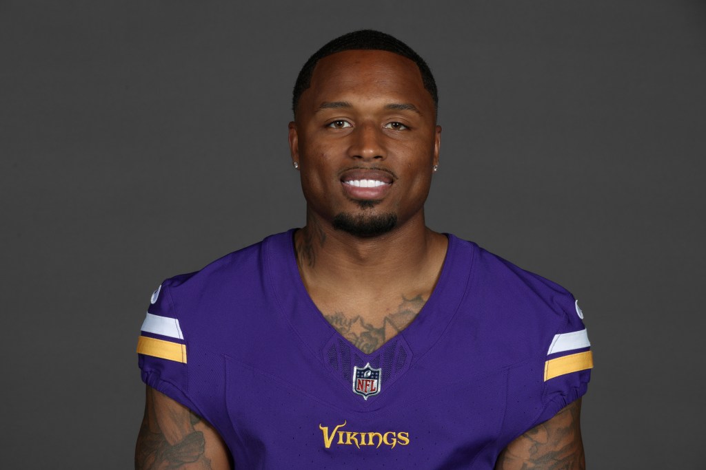 Portrait of a male football player wearing a purple NFL jersey, smiling against a gray background.