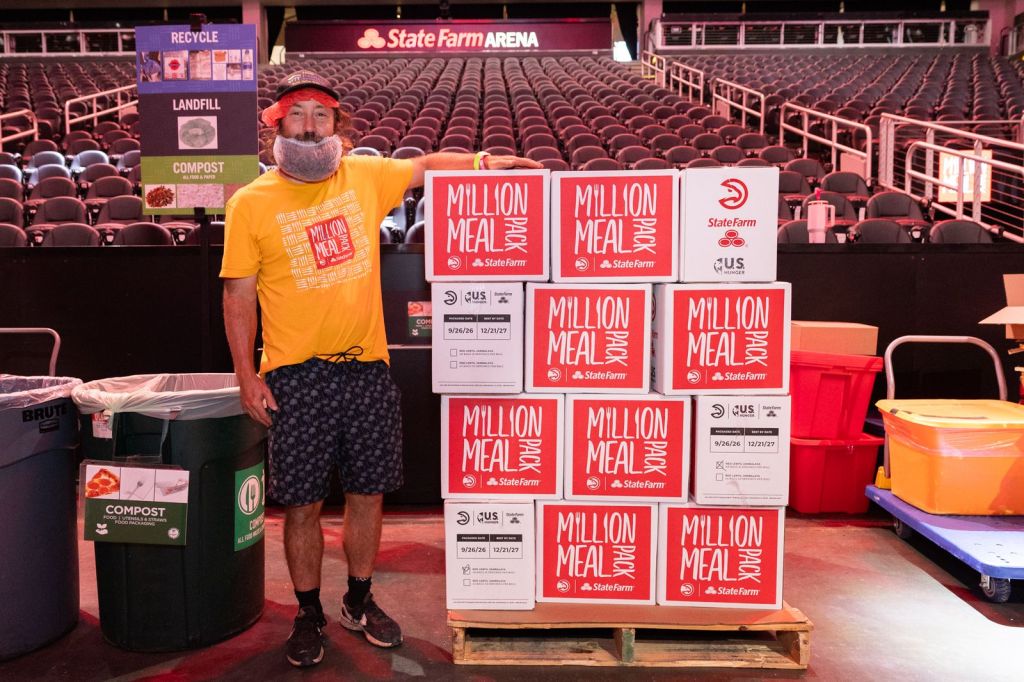 A volunteer posing next to a stack of food boxes labeled 'MILLION MEAL PACK' inside a sports arena, with recycling, landfill, and compost bins in the background.