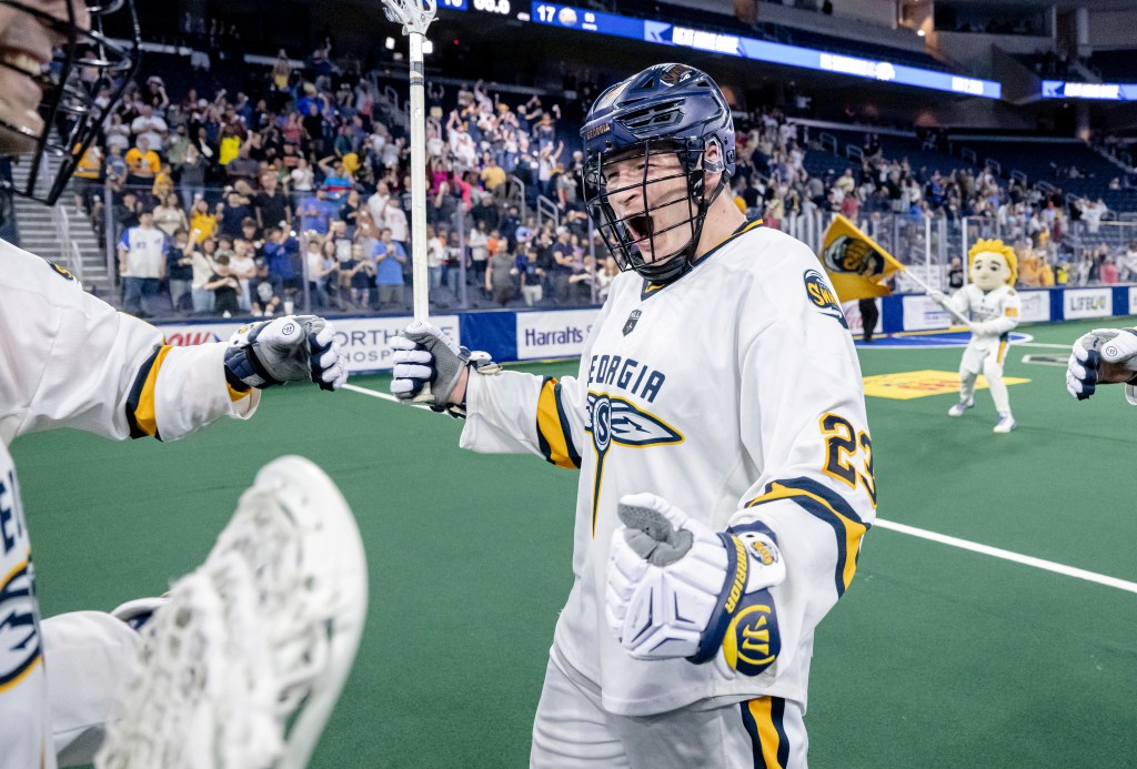 A lacrosse player celebrating on the field with teammates in a packed arena, wearing a white jersey with blue and yellow accents, and a helmet. The crowd in the background is cheering, and a team mascot is visible.