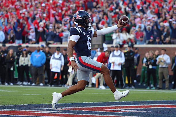 A football player in a blue and red uniform celebrating after scoring a touchdown, with fans in the background.