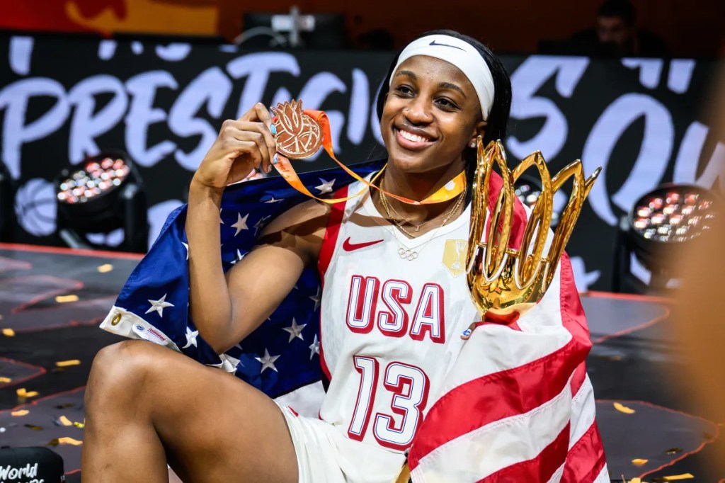 Smiling athlete in a basketball uniform holding a trophy and wearing a medal, wrapped in an American flag.