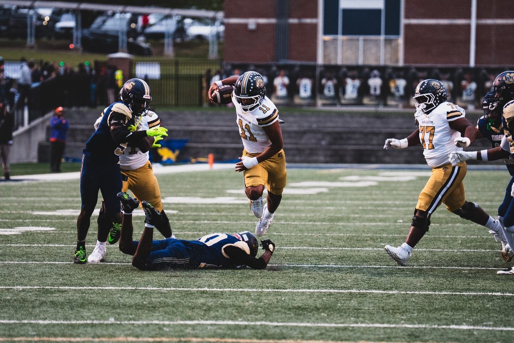 A football player in a white uniform leaps over a downed opponent on the field during a game, with other players in the background.
