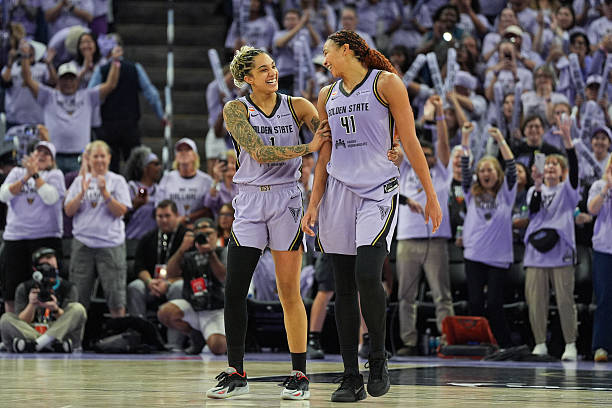 Two basketball players smiling and celebrating on the court, with a cheering crowd in the background.