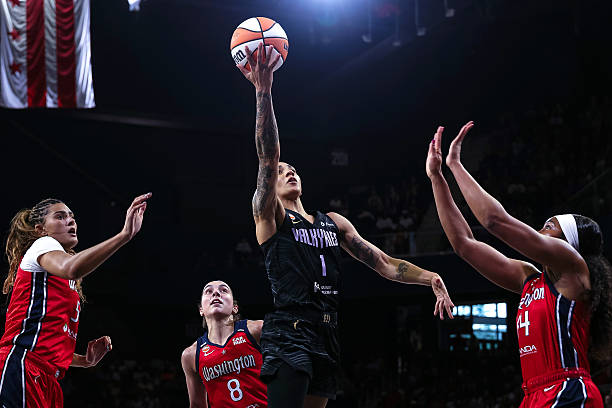 A basketball player in a black uniform leaps to make a shot while being defended by three players in red uniforms, showcasing athleticism and competition in a dynamic game setting.