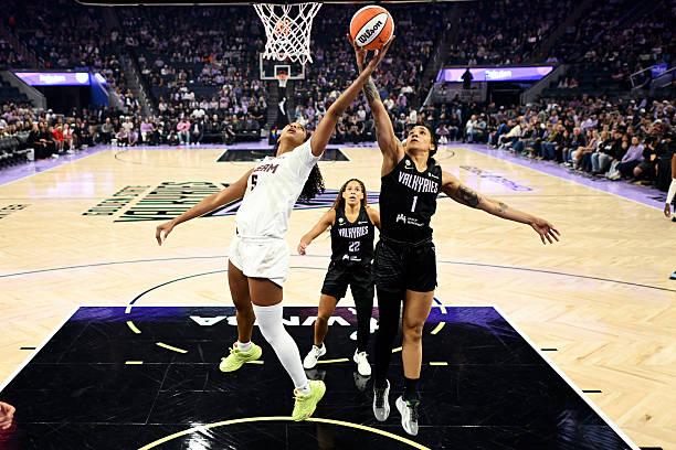 A basketball player leaps to block a shot during a game, with another player in white attempting to score. The crowd is visible in the background.