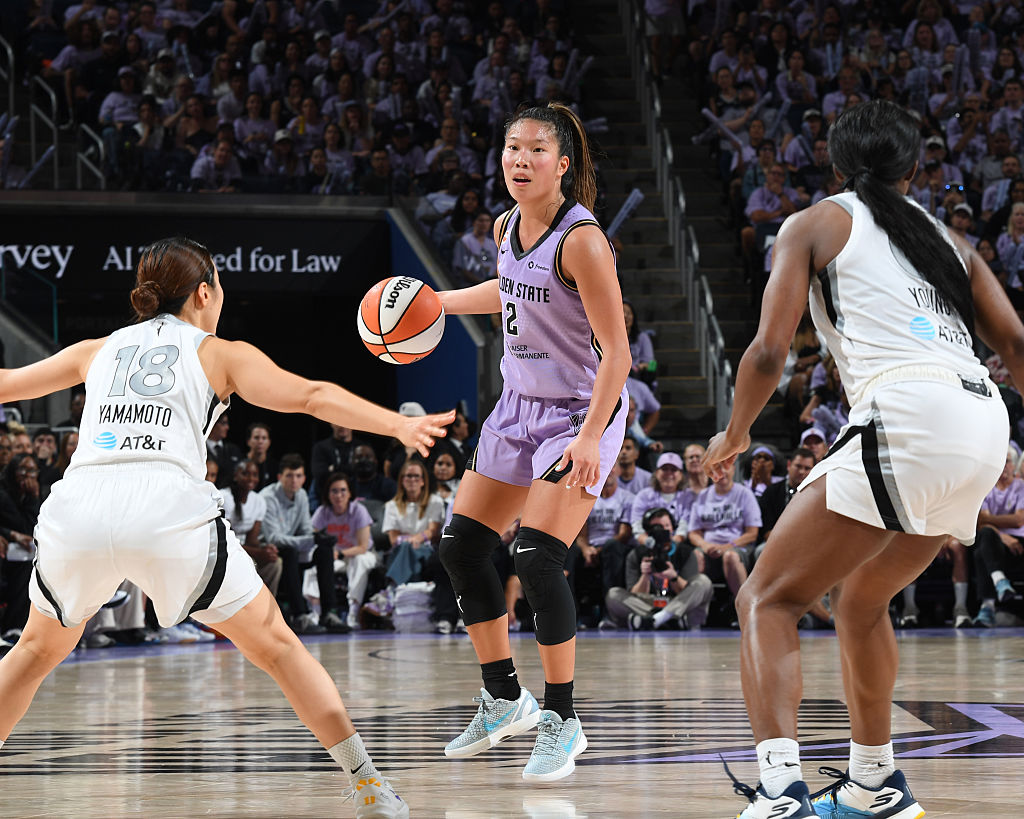 A basketball player wearing a purple jersey with the number 2 is preparing to pass the ball during a game, while two defenders in white jerseys, one of whom has the number 18, are positioning themselves to intercept.