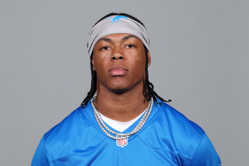 A male athlete wearing a blue NFL jersey with a gray headband and multiple necklaces, posing against a gray background.