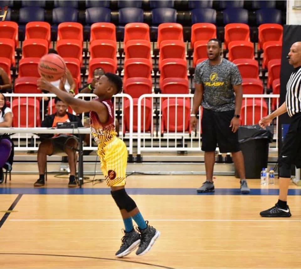 A young boy in a colorful basketball uniform jumps to shoot a basketball during a game, with spectators and coaches visible in the background.