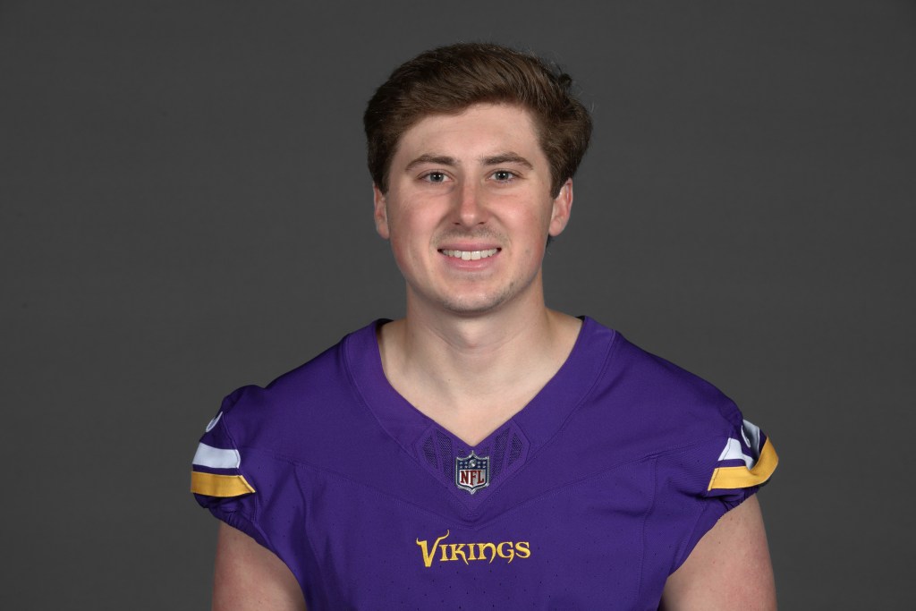 Portrait of a smiling male football player wearing a purple Minnesota Vikings jersey with NFL logo on a plain gray background.