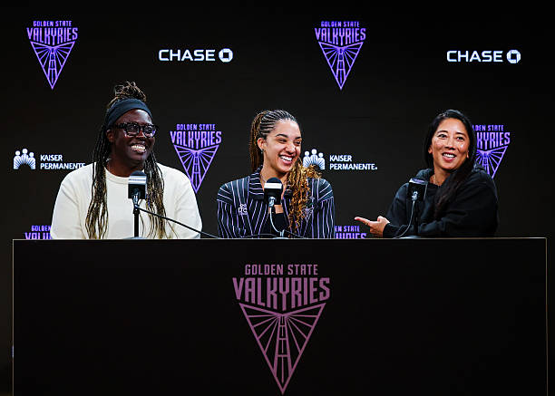 Three women sitting at a press conference table with microphones, smiling and interacting. The backdrop features the logo for the Golden State Valkyries and sponsor logos.