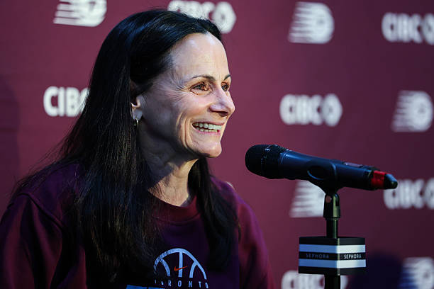 A woman with long dark hair smiling while sitting at a microphone during a press conference, wearing a maroon shirt with a basketball logo, against a branded backdrop.