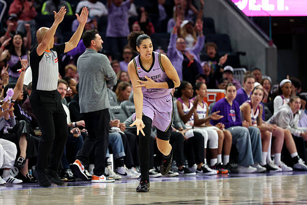 A basketball player in a purple uniform celebrates a play during a game, while teammates and fans cheer from the sidelines.