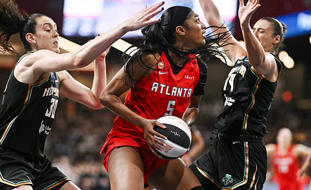 Action shot of a female basketball player in a red Atlanta jersey, driving towards the basket while being challenged by two defenders in black jerseys.