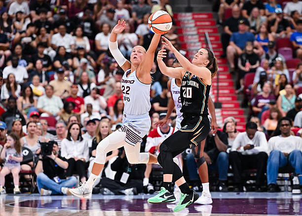 A female basketball player in a white jersey reaching for a ball while another player in a black jersey defends, with a crowd in the background.