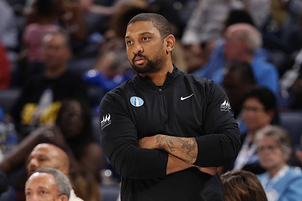 A man wearing a black athletic jacket with his arms crossed, watching a basketball game from the sidelines.