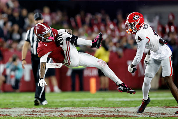 A college football player from Alabama is tackled while running the ball, with a player from Georgia in pursuit. The scene captures the intensity of the game with referees in the background.