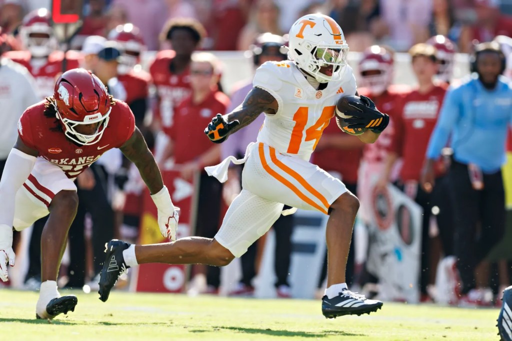 A football player in a white and orange uniform is running with the ball while evading a defender in a red uniform during a game.