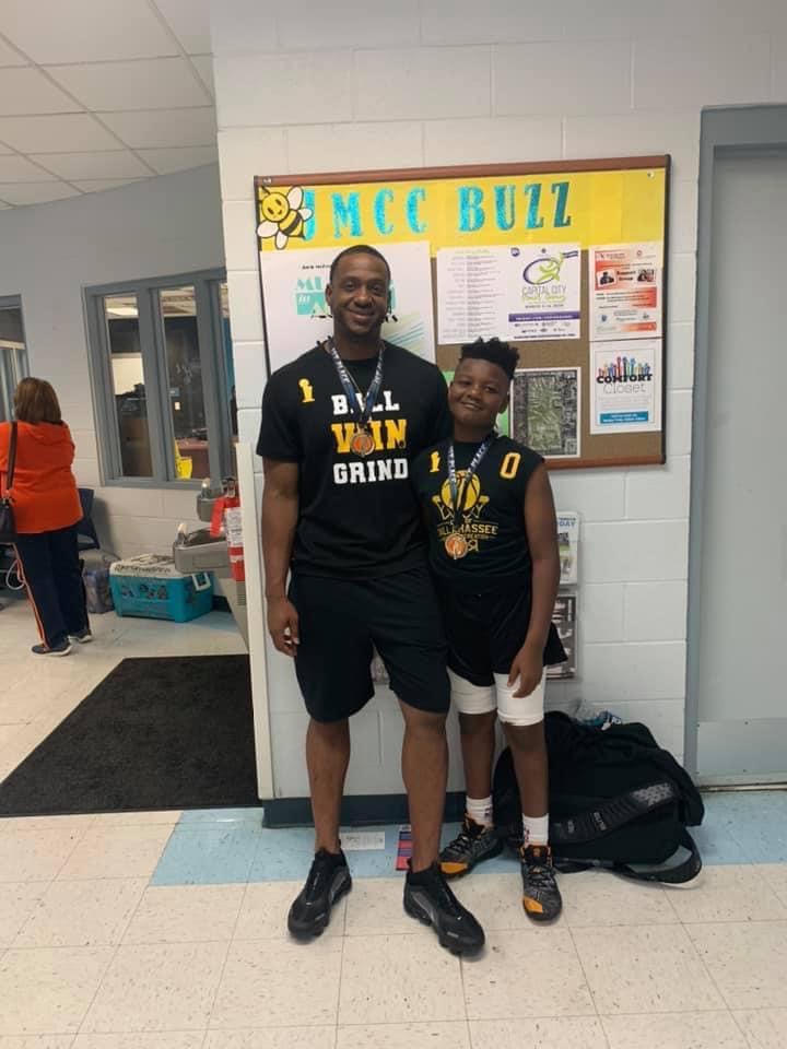 A man and a boy pose together inside a gymnasium, both wearing basketball uniforms and medals. The man is wearing a black t-shirt that reads 'BALL VON GRIND', while the boy is smiling beside him. In the background, there's a bulletin board with various announcements and information.